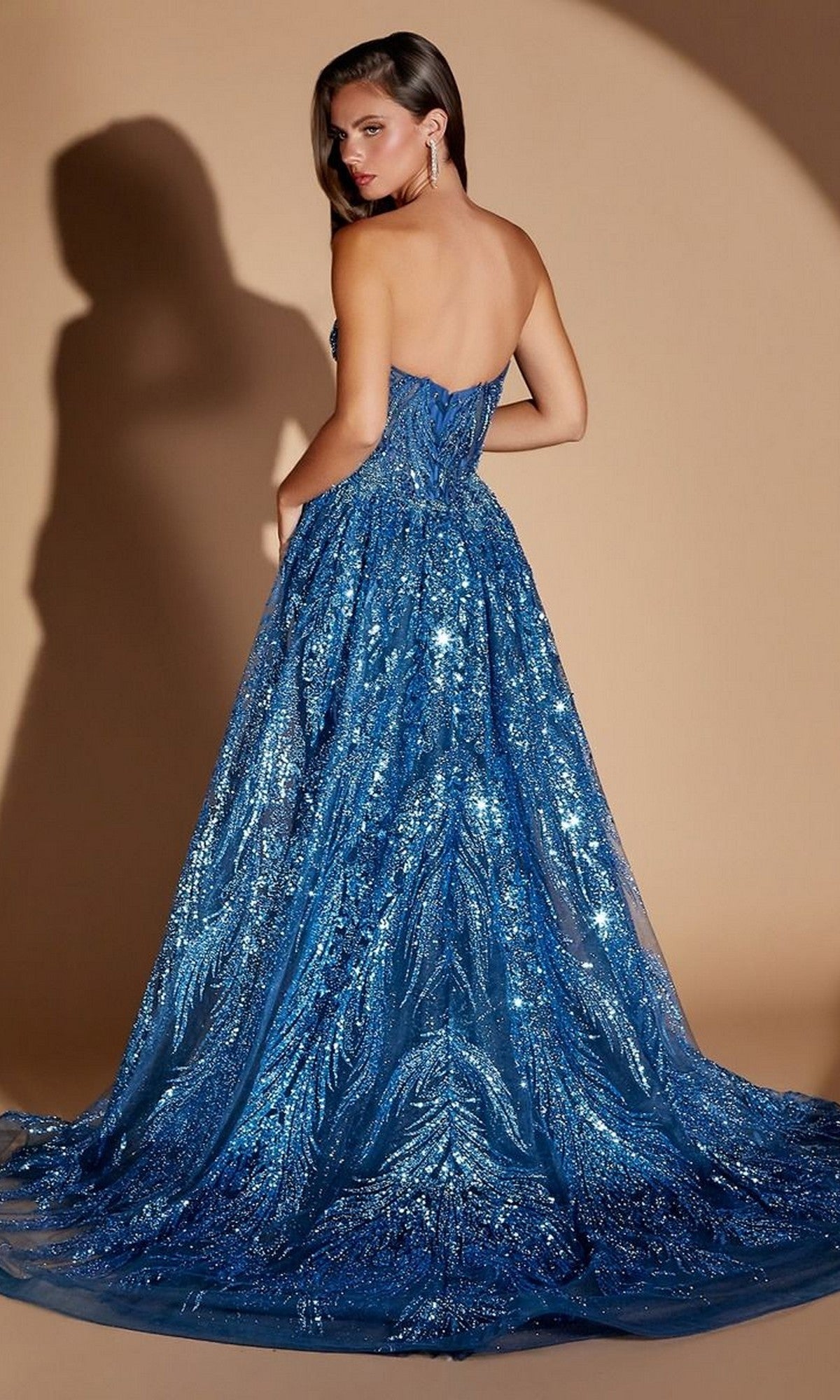 A woman wears the Ladivine CM393 Long Prom Dress—a strapless, floor-length blue gown embellished with glitter—standing with her back slightly turned and looking over her shoulder against a beige background.